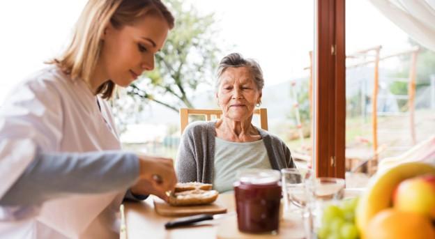 An older adult watching a caregiver get breakfast ready at the table in an assisted living community.